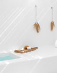 KULALA Living minimalist white bathroom featuring natural raffia and shell hanging decor, a wooden bath tray with soap and sponge, and a tranquil built-in bath