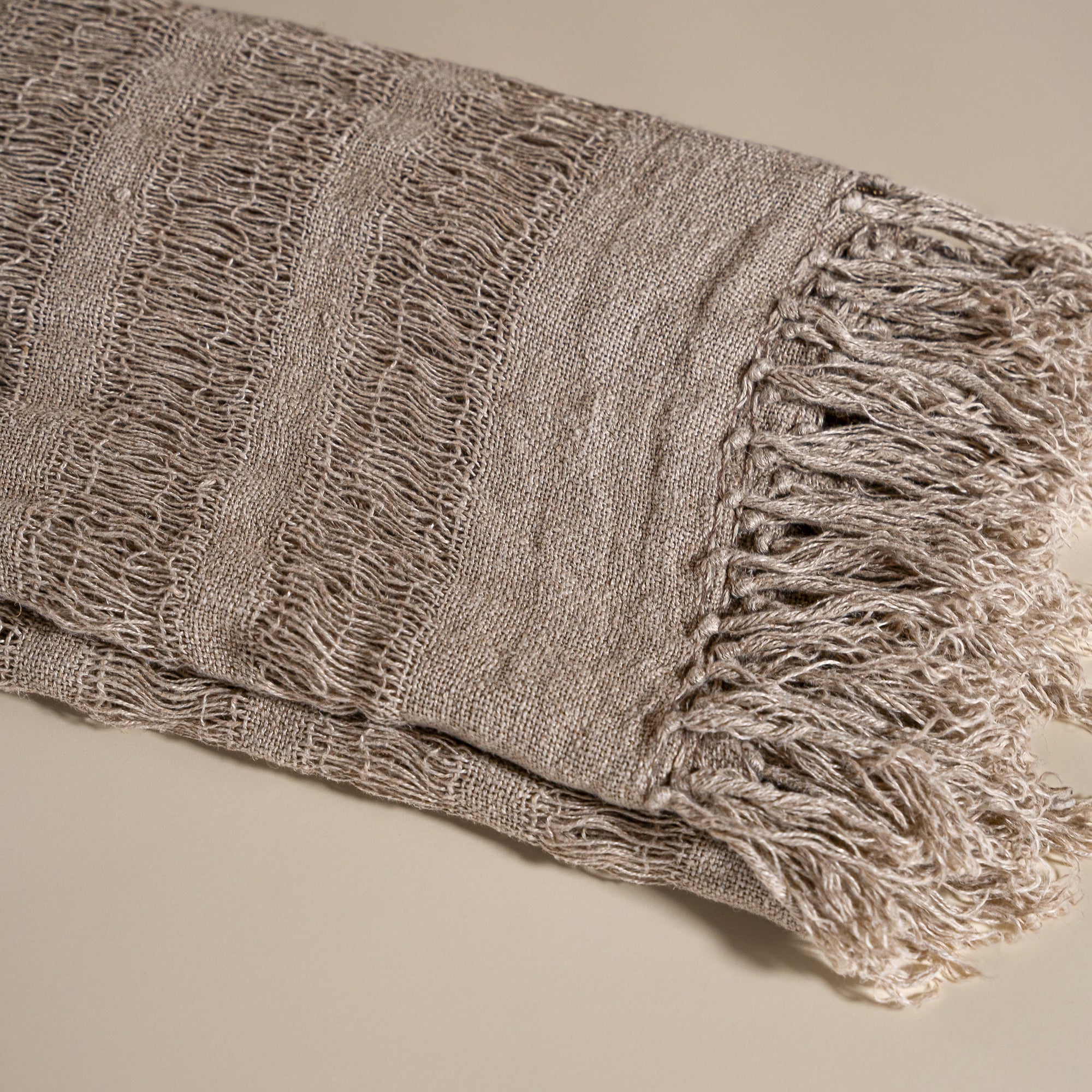 Handwoven linen throw in earthy taupe tones, neatly folded to highlight its delicate open-weave texture and soft fringe detail, placed on a neutral background