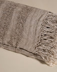 Handwoven linen throw in earthy taupe tones, neatly folded to highlight its delicate open-weave texture and soft fringe detail, placed on a neutral background