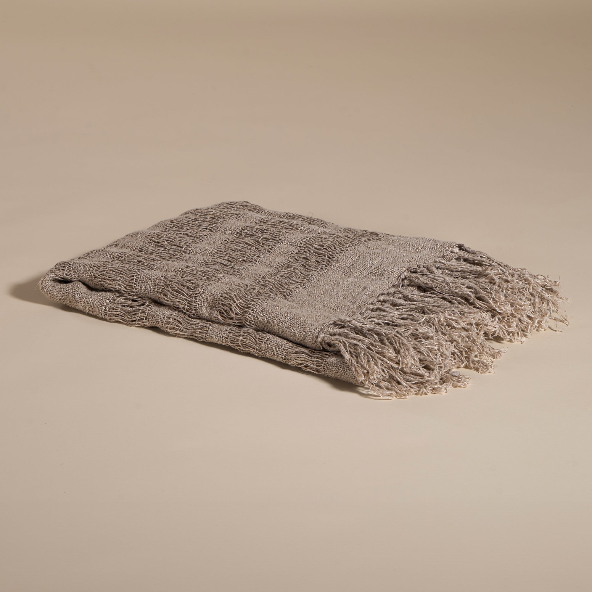 Handwoven linen throw in earthy taupe tones, neatly folded to highlight its delicate open-weave texture and soft fringe detail, placed on a neutral background