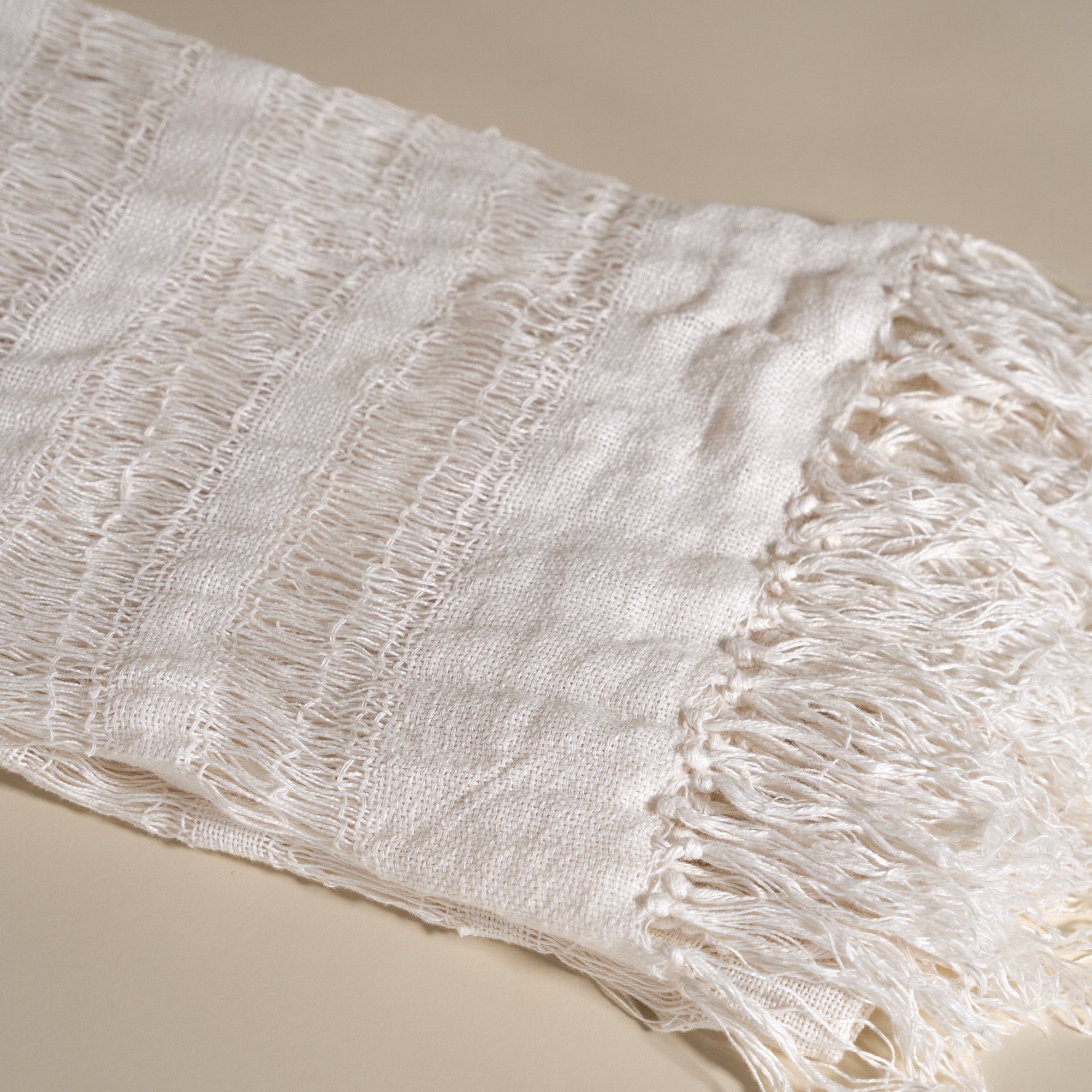 Handwoven linen throw in neutral tones, neatly folded to highlight its delicate open-weave texture and soft fringe detail, placed on a neutral background