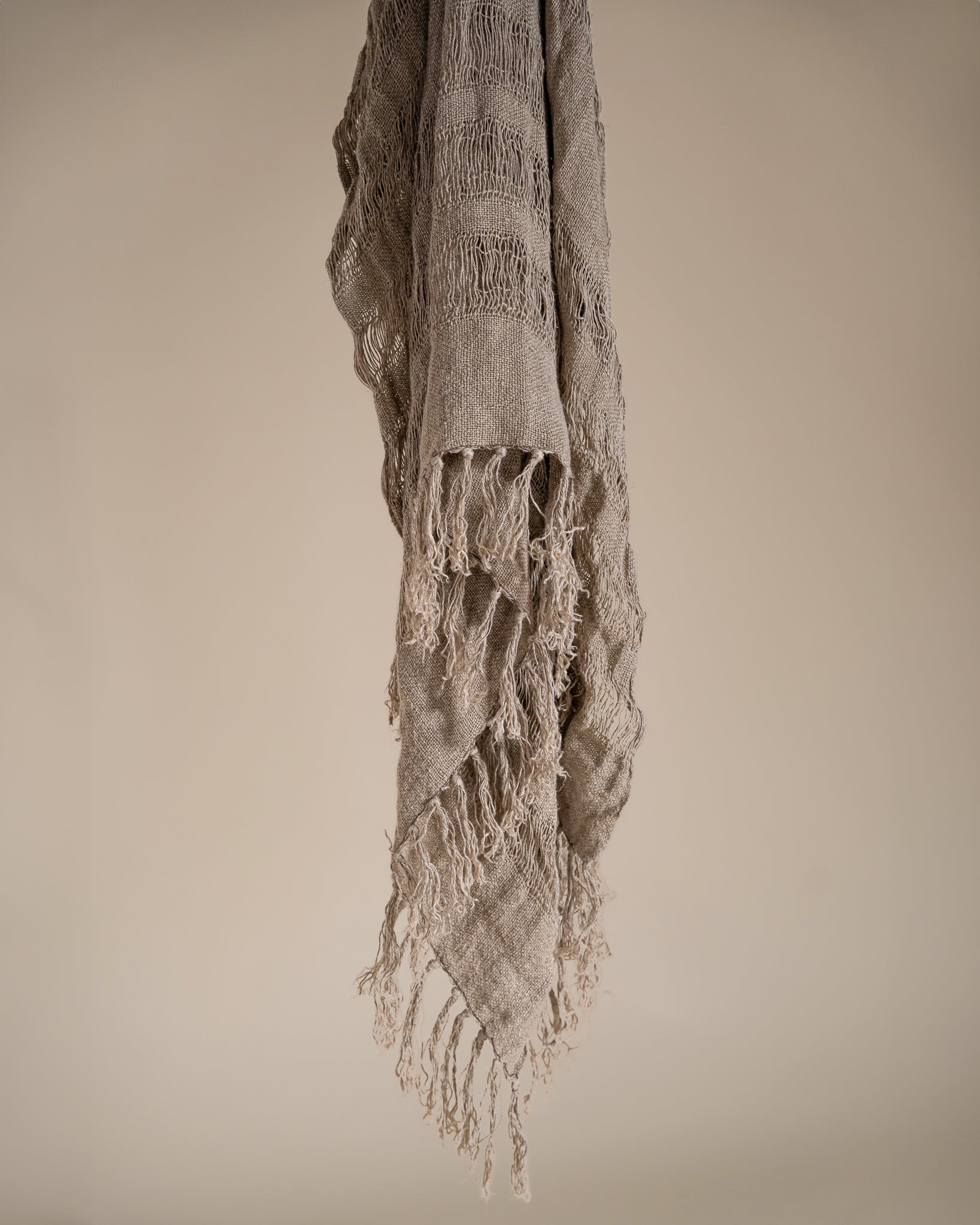 Handwoven natural linen throw in a soft neutral tone, featuring an intricate open-weave pattern and delicate fringed edges, elegantly draped against a beige backdrop