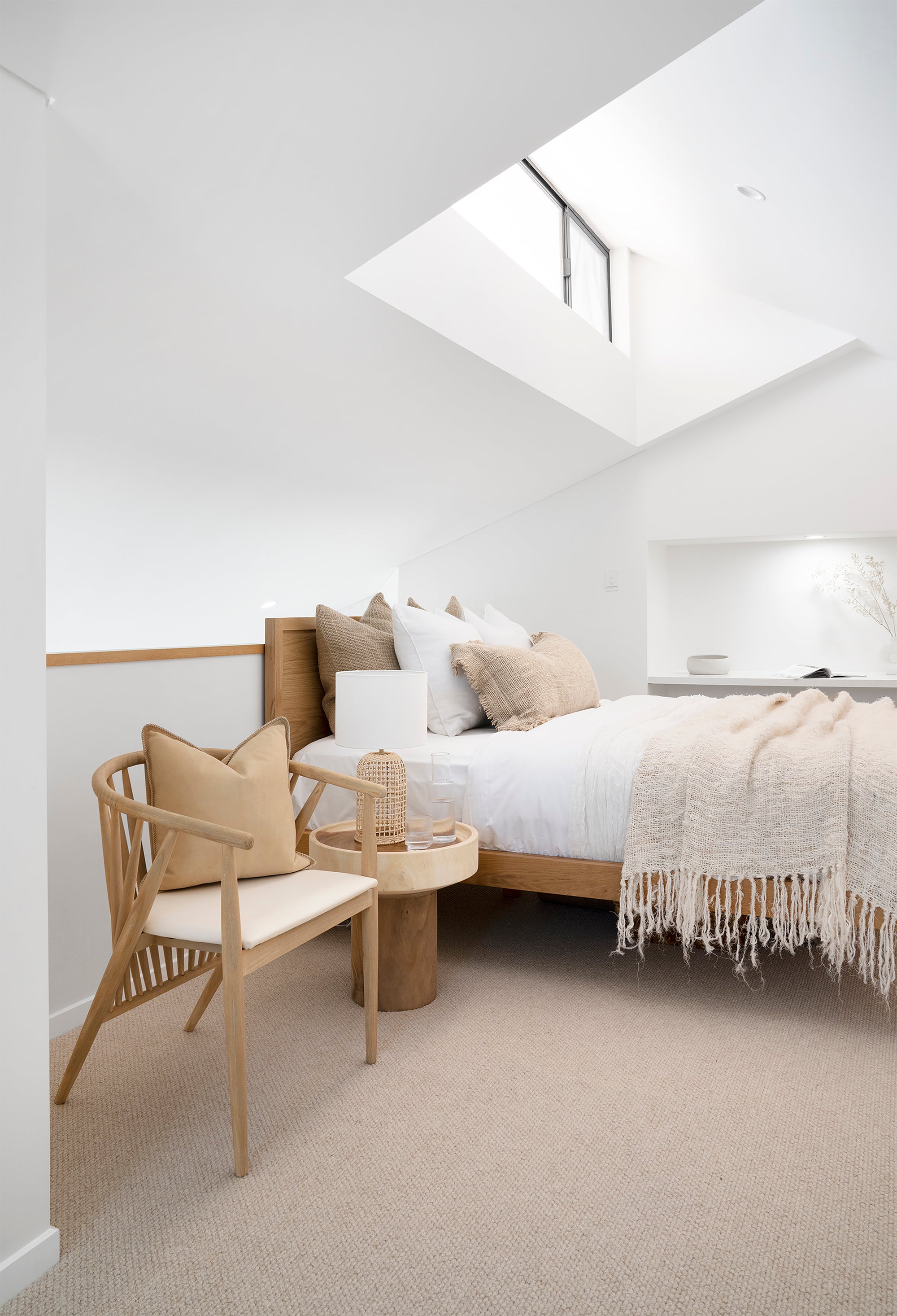 Minimalist loft bedroom featuring a timber bed frame with layered white and beige linen, textured cushions, and a soft woven throw. A lightwood spindle accent chair with a neutral cushion sits beside a round wooden side table with a woven lamp, enhancing the serene, natural aesthetic