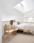 Minimalist loft bedroom featuring a timber bed frame with layered white and beige linen, textured cushions, and a soft woven throw. A lightwood spindle accent chair with a neutral cushion sits beside a round wooden side table with a woven lamp, enhancing the serene, natural aesthetic