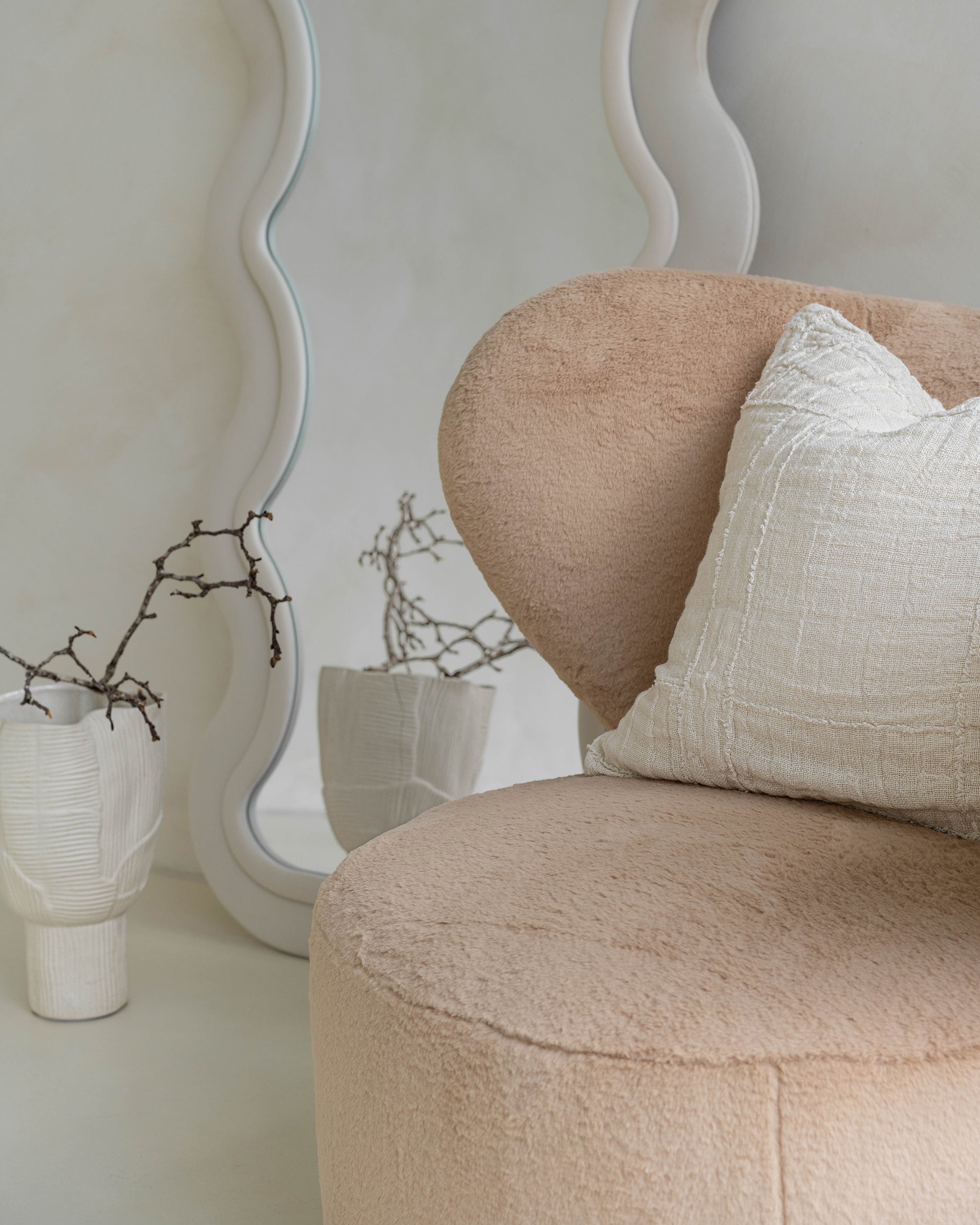 Modern lounge corner featuring a beige teddy armchair with a textured throw and linen cushion, styled beside a sculptural wavy mirror. Neutral-toned ceramics and organic branches complete the serene, minimalist aesthetic