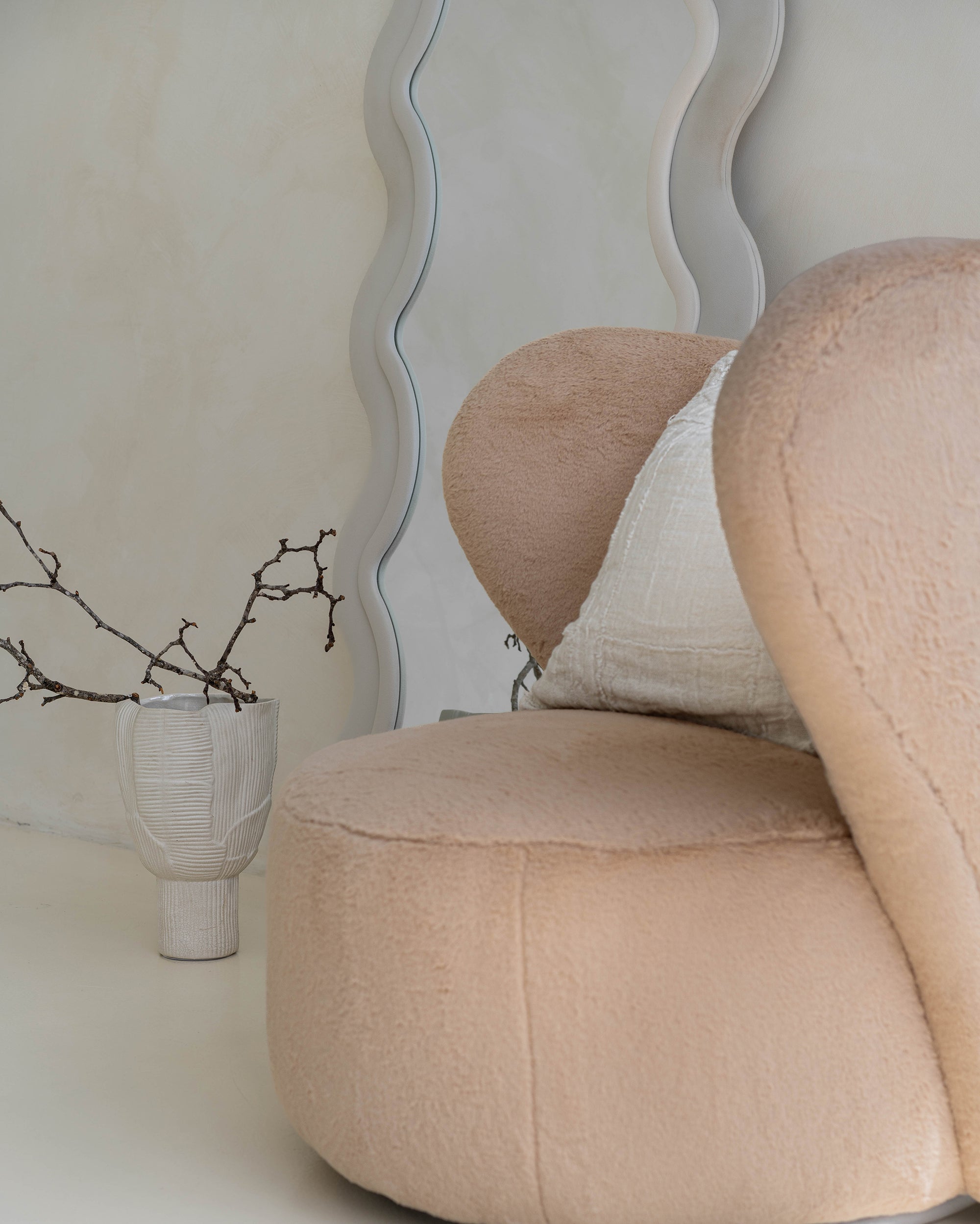 Modern lounge corner featuring a beige teddy armchair with a linen cushion, styled beside a sculptural wavy mirror. Neutral-toned ceramics and organic branches complete the serene, minimalist aesthetic