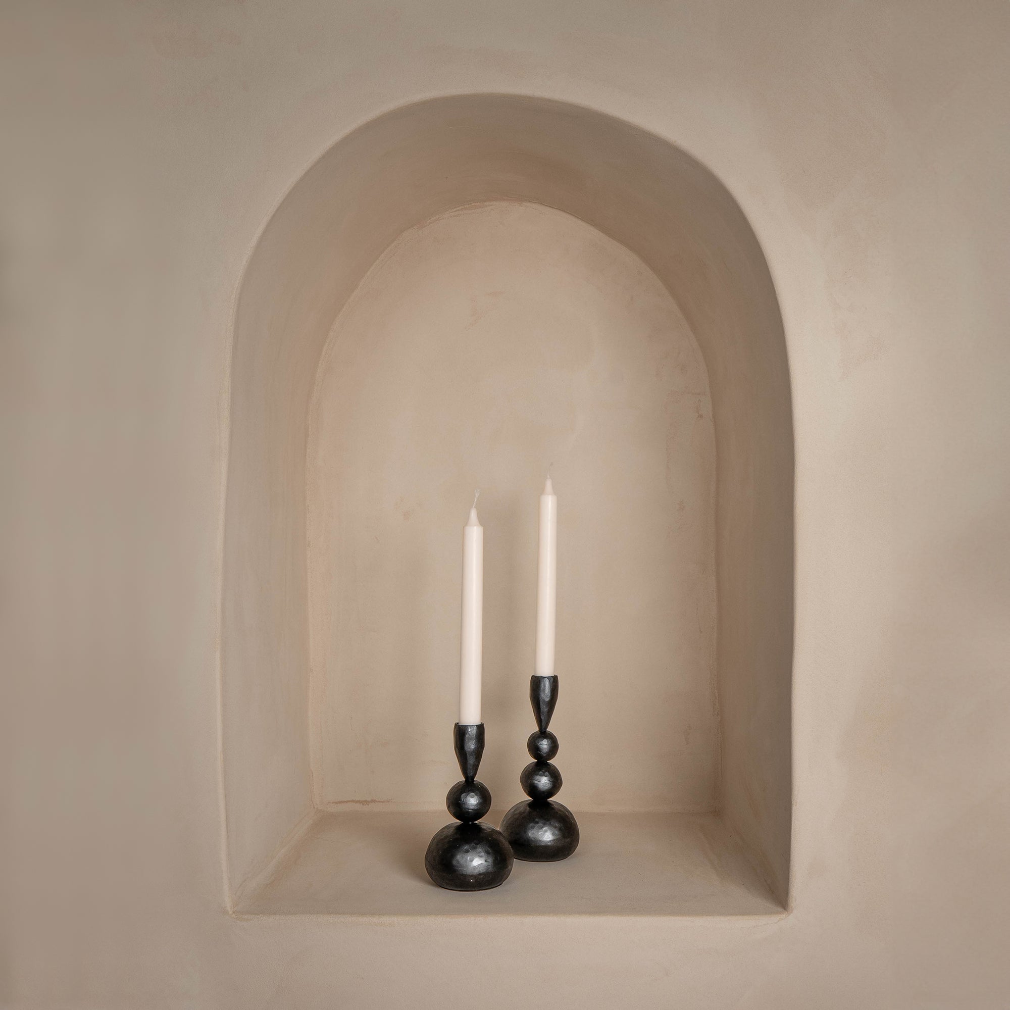 A pair of sculptural black iron candle holders with an organic, modern design, elegantly placed in a soft beige arched wall niche, creating a warm and minimalist aesthetic