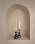 A pair of sculptural black iron candle holders with an organic, modern design, elegantly placed in a soft beige arched wall niche, creating a warm and minimalist aesthetic