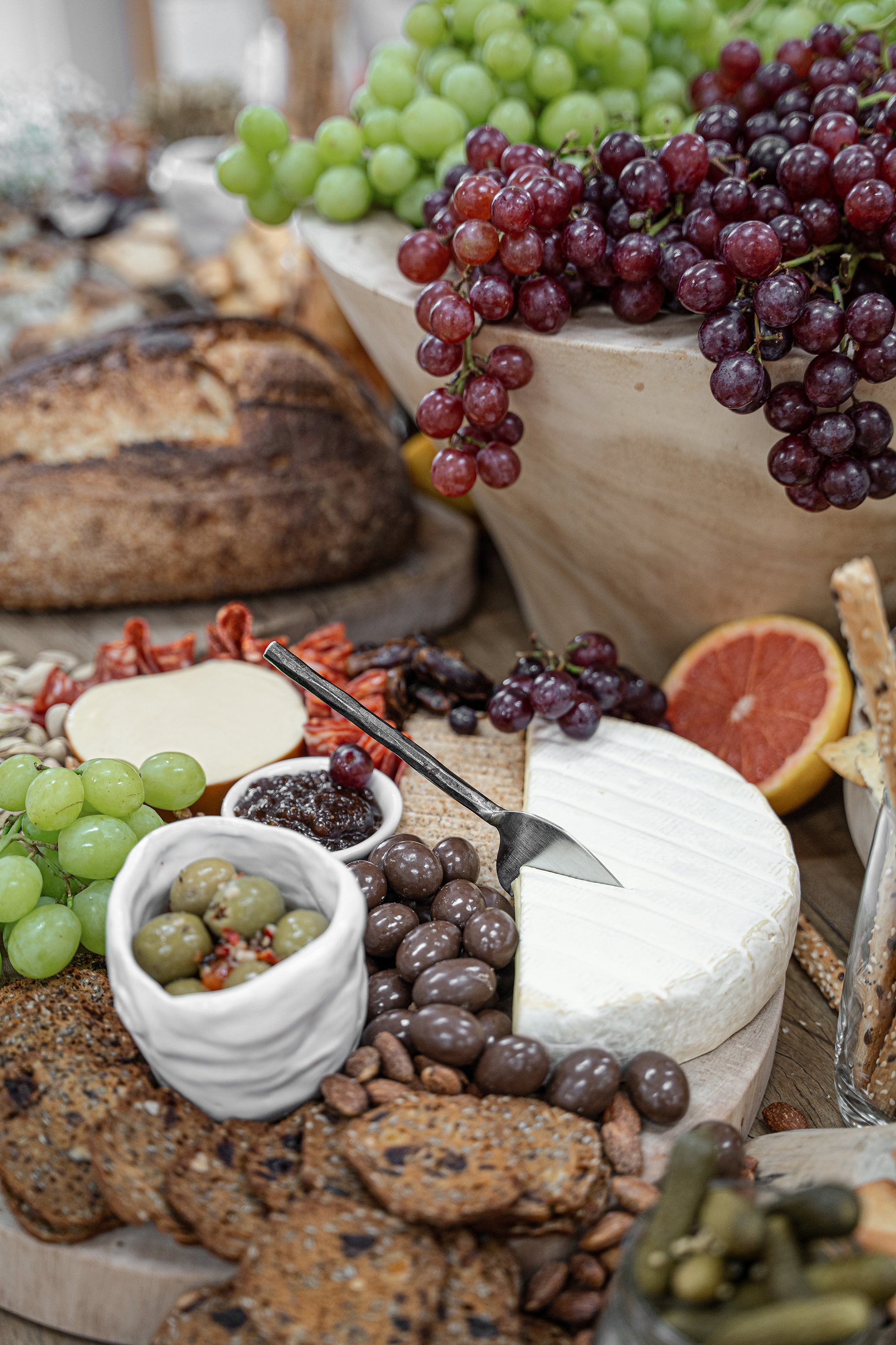 A luxurious grazing board featuring KULALA Living’s black hand-forged cheese knife cutting into a wheel of creamy brie. Surrounded by crisp crackers, rich chocolate-covered nuts, olives, and fresh grapes, this setting is perfect for effortless entertaining