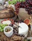 A luxurious grazing board featuring KULALA Living’s black hand-forged cheese knife cutting into a wheel of creamy brie. Surrounded by crisp crackers, rich chocolate-covered nuts, olives, and fresh grapes, this setting is perfect for effortless entertaining