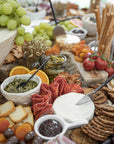 An abundant grazing board featuring KULALA Living’s black hand-forged cheese knife slicing through creamy brie. Surrounded by gourmet crackers, cured meats, fresh fruit, and savoury dips, this scene embodies effortless, elevated entertaining