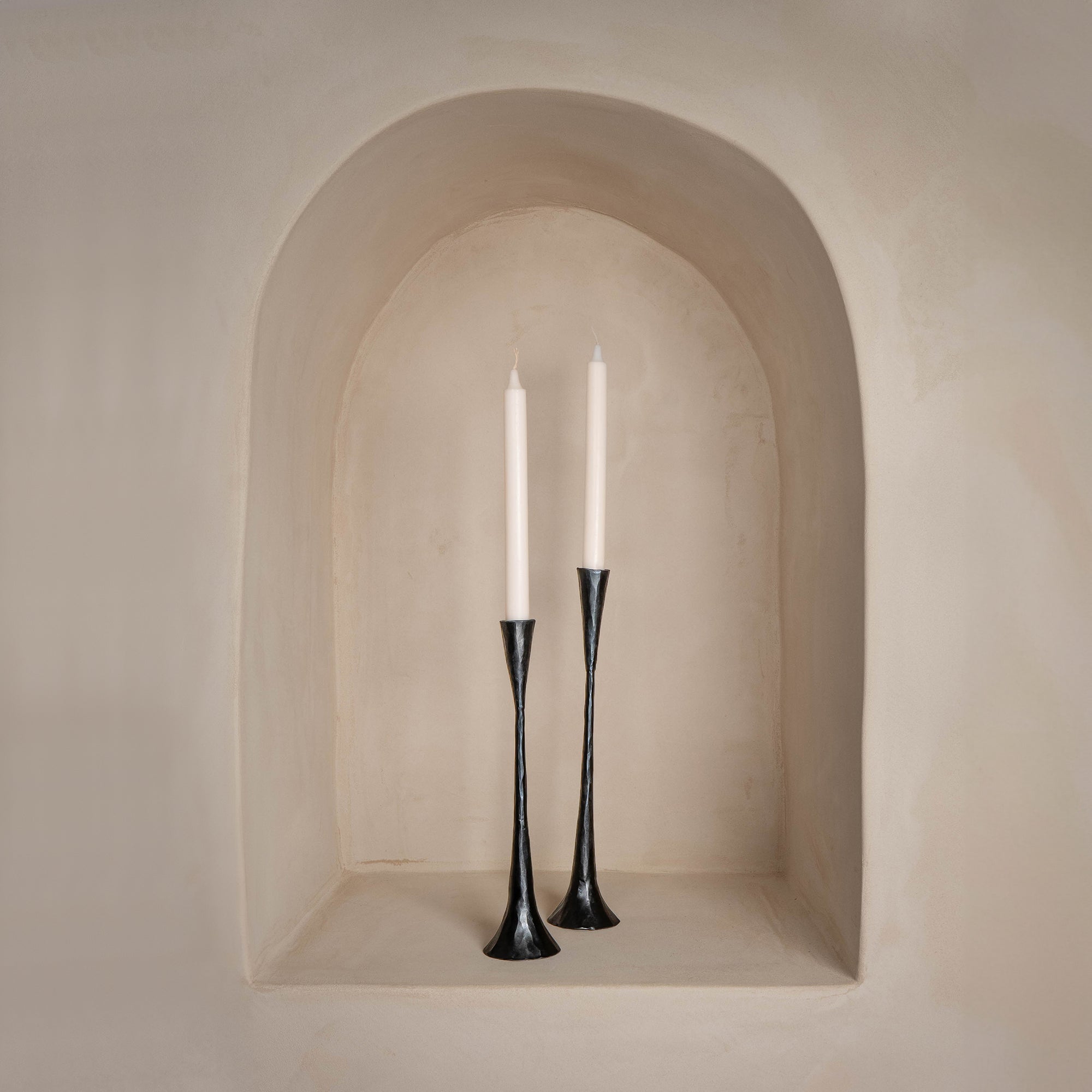 A pair of slender black metal taper candle holders with a sculptural, elongated design, displayed in a softly curved alcove with a warm neutral-toned backdrop