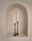 A pair of slender black metal taper candle holders with a sculptural, elongated design, displayed in a softly curved alcove with a warm neutral-toned backdrop