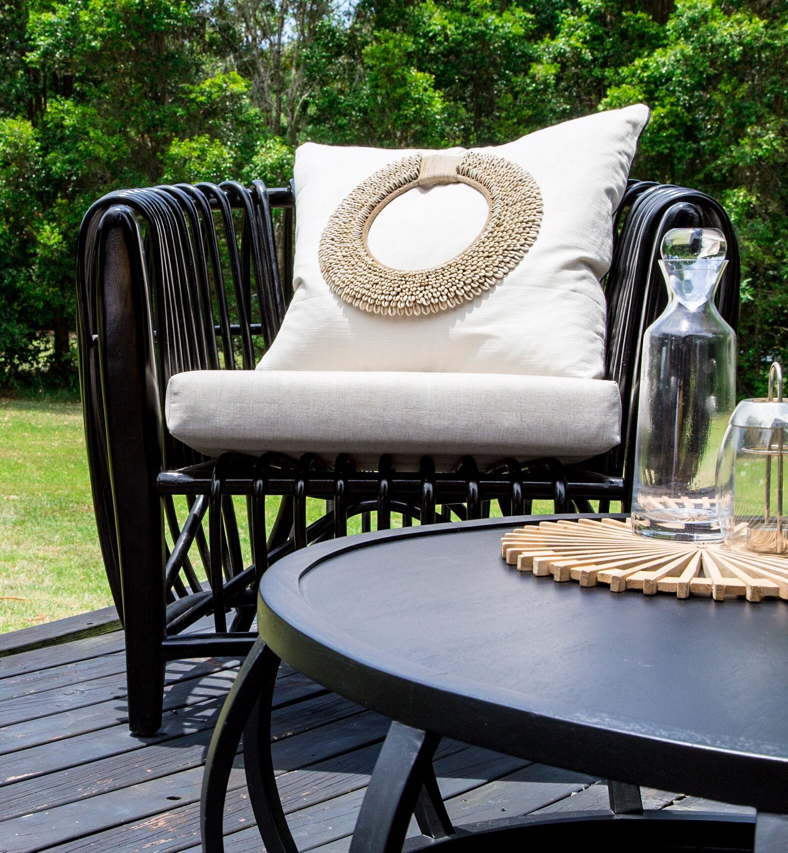 Black rattan lounge chair with neutral cushion and decorative shell pillow, styled on an outdoor deck with a black round coffee table and glassware