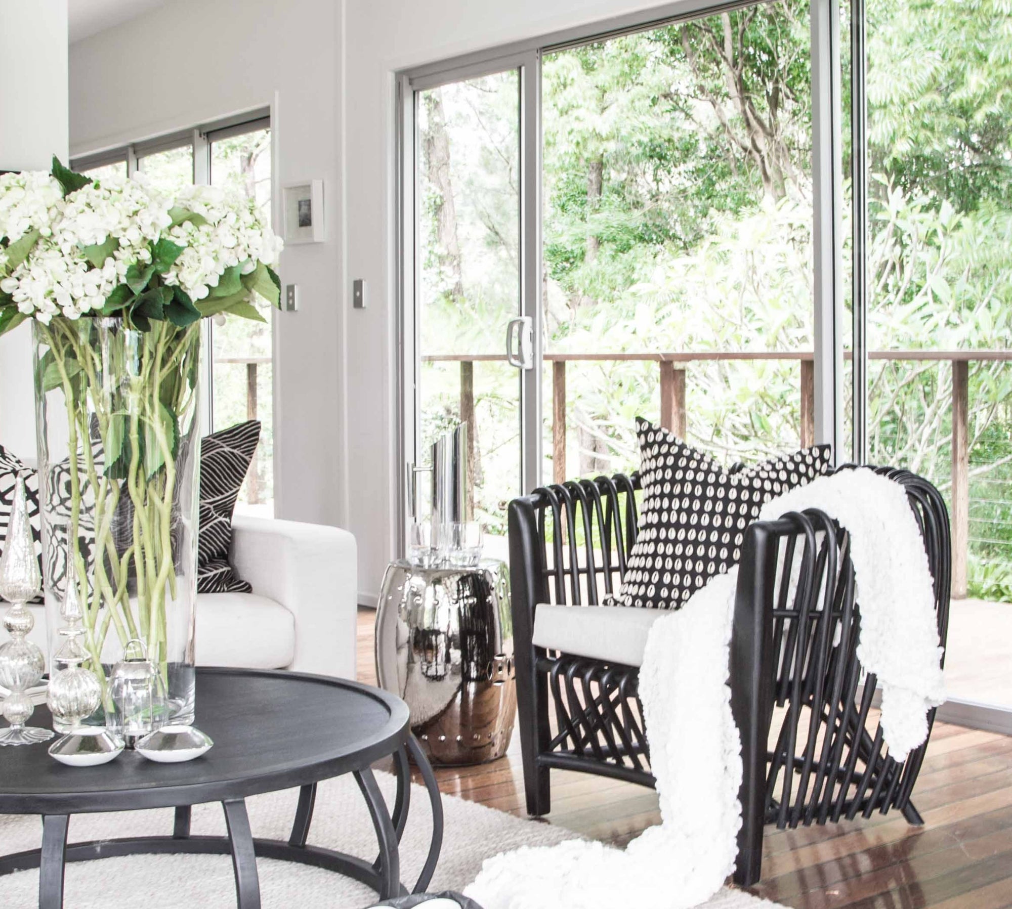 Modern living room featuring a black rattan lounge chair with a neutral cushion, accented with monochrome decor and lush greenery views