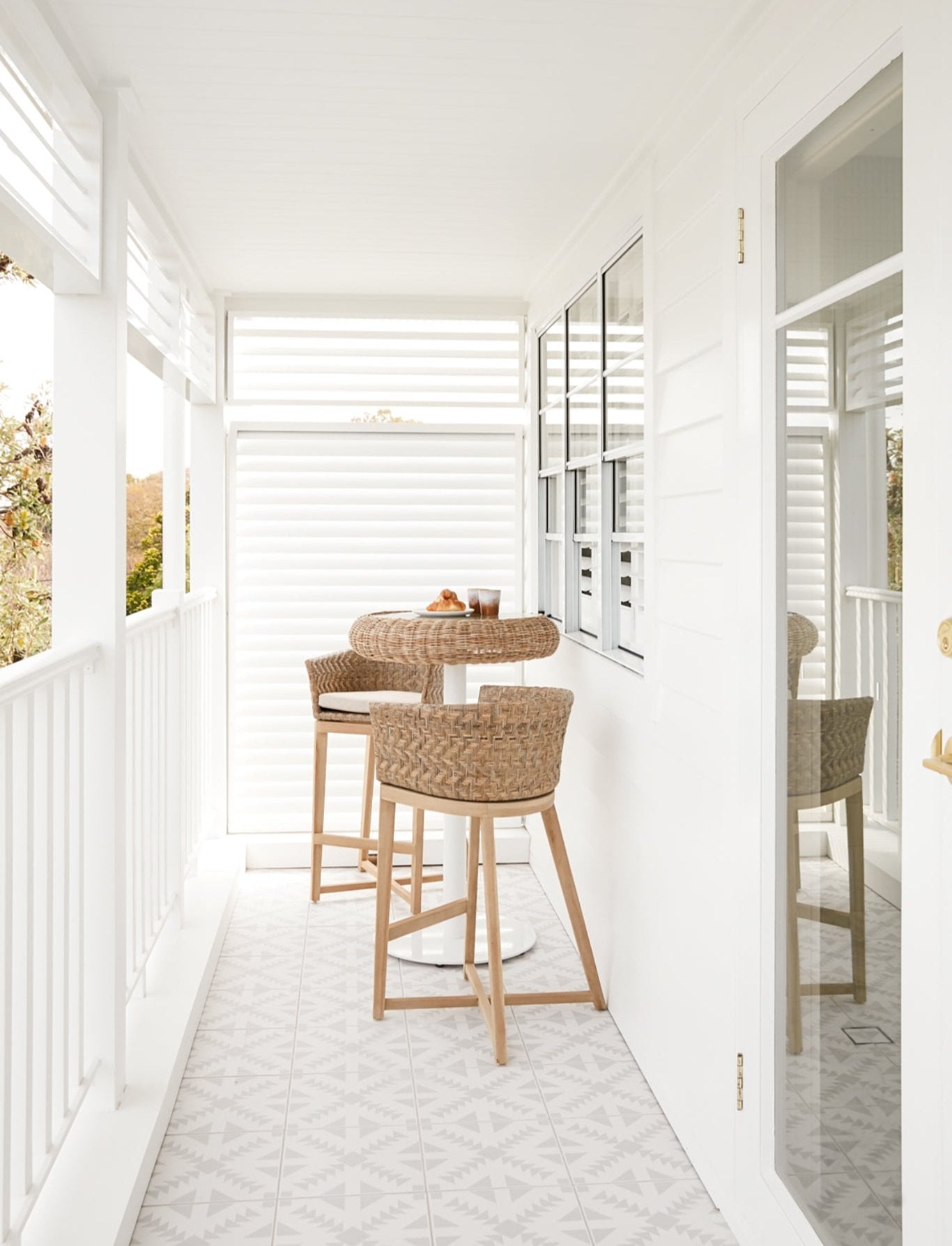 Coastal-inspired outdoor bistro setting featuring woven rattan bar stools with cushioned seats and a glass-top rattan dining table, styled with fresh croissants and coffee