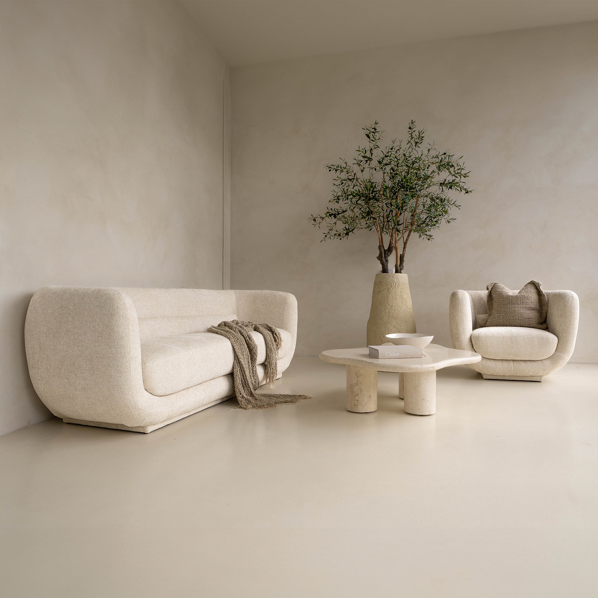 A modern, minimalist lounge setting featuring the KULALA Living curved bouclé sofa and matching armchair in a soft ivory hue. Styled with a textured throw, linen cushions, a sculptural stone coffee table, and a statement vase with an olive tree for a tranquil, contemporary aesthetic