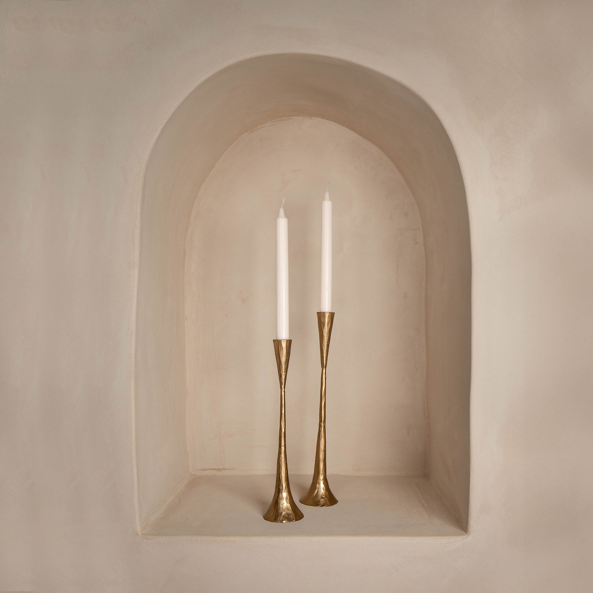 A pair of tall brass candlesticks with a hammered finish, holding white taper candles, elegantly displayed in an arched wall niche with a smooth, neutral-toned plaster finish