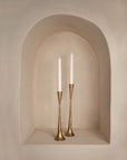 A pair of tall brass candlesticks with a hammered finish, holding white taper candles, elegantly displayed in an arched wall niche with a smooth, neutral-toned plaster finish