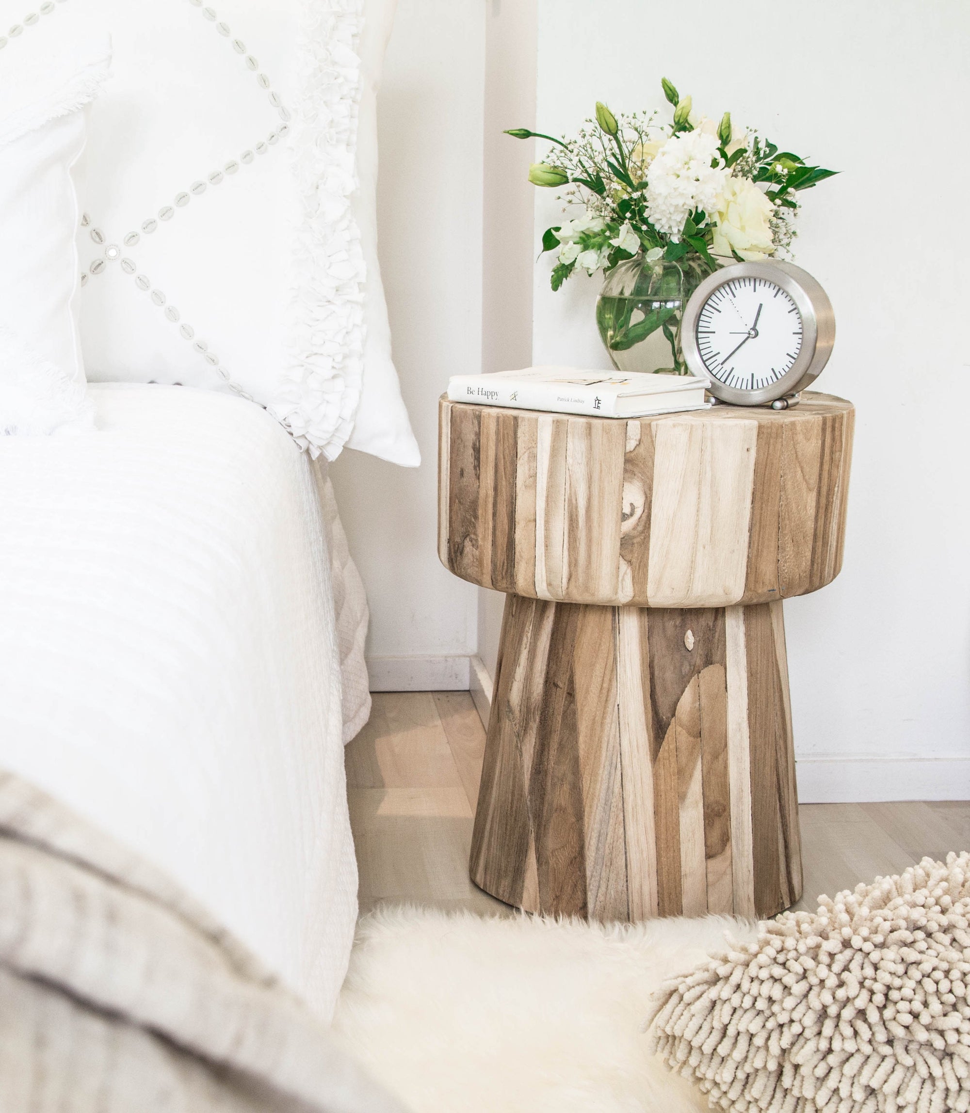 Serene bedroom featuring a natural teak side table styled with fresh flowers, books, and a minimalist clock, complementing the soft white bedding and cosy textures.