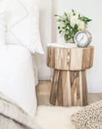 Serene bedroom featuring a natural teak side table styled with fresh flowers, books, and a minimalist clock, complementing the soft white bedding and cosy textures.