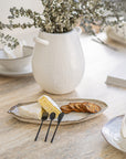 A textured white ceramic vase with dried foliage, styled on a travertine table alongside a rustic ceramic cheese platter with brie, crackers, and black serving cutlery