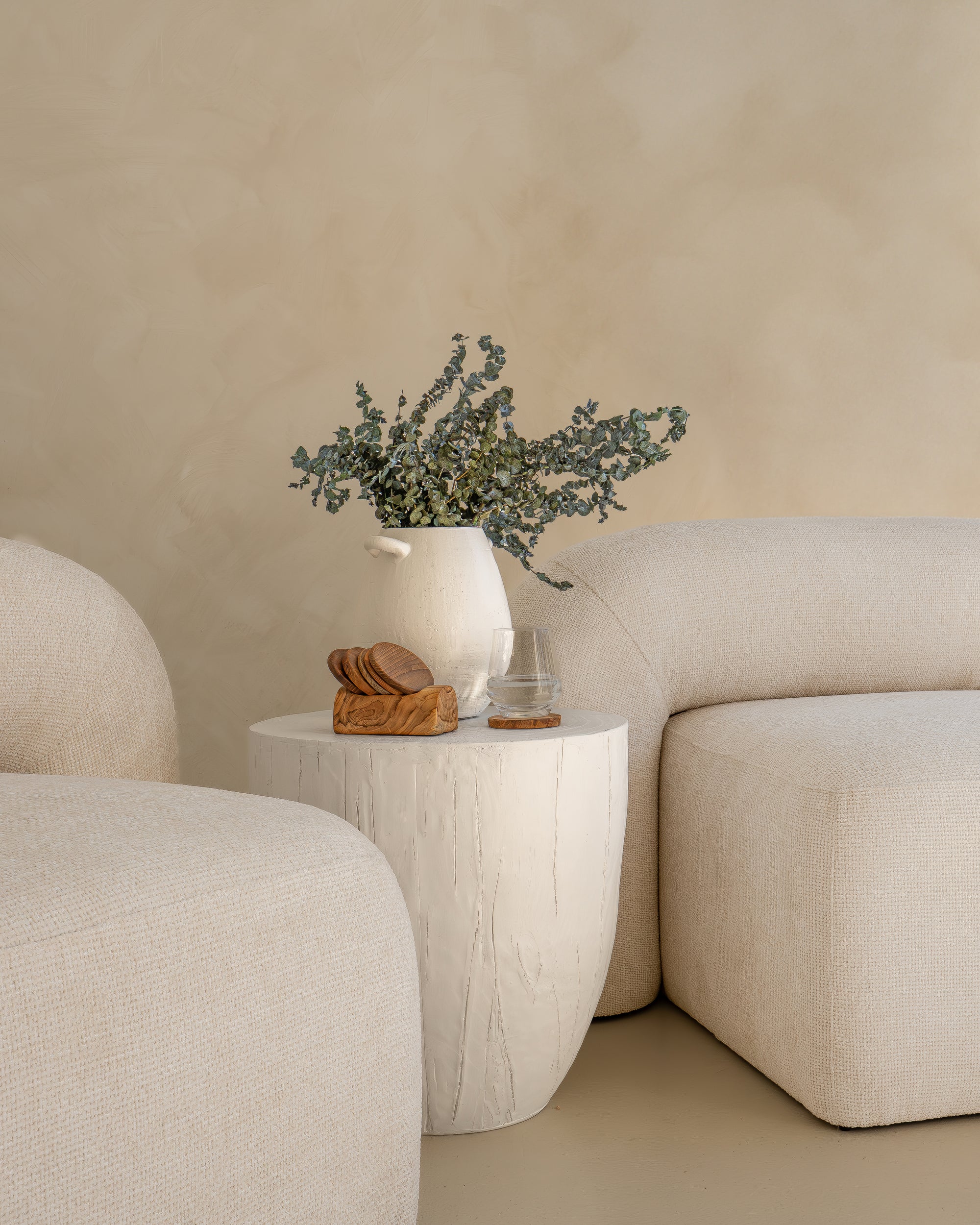 A white ceramic vase with dried foliage sits atop a textured side table, styled with a wooden coaster, glassware, and crackers, nestled between plush curved cream sofas