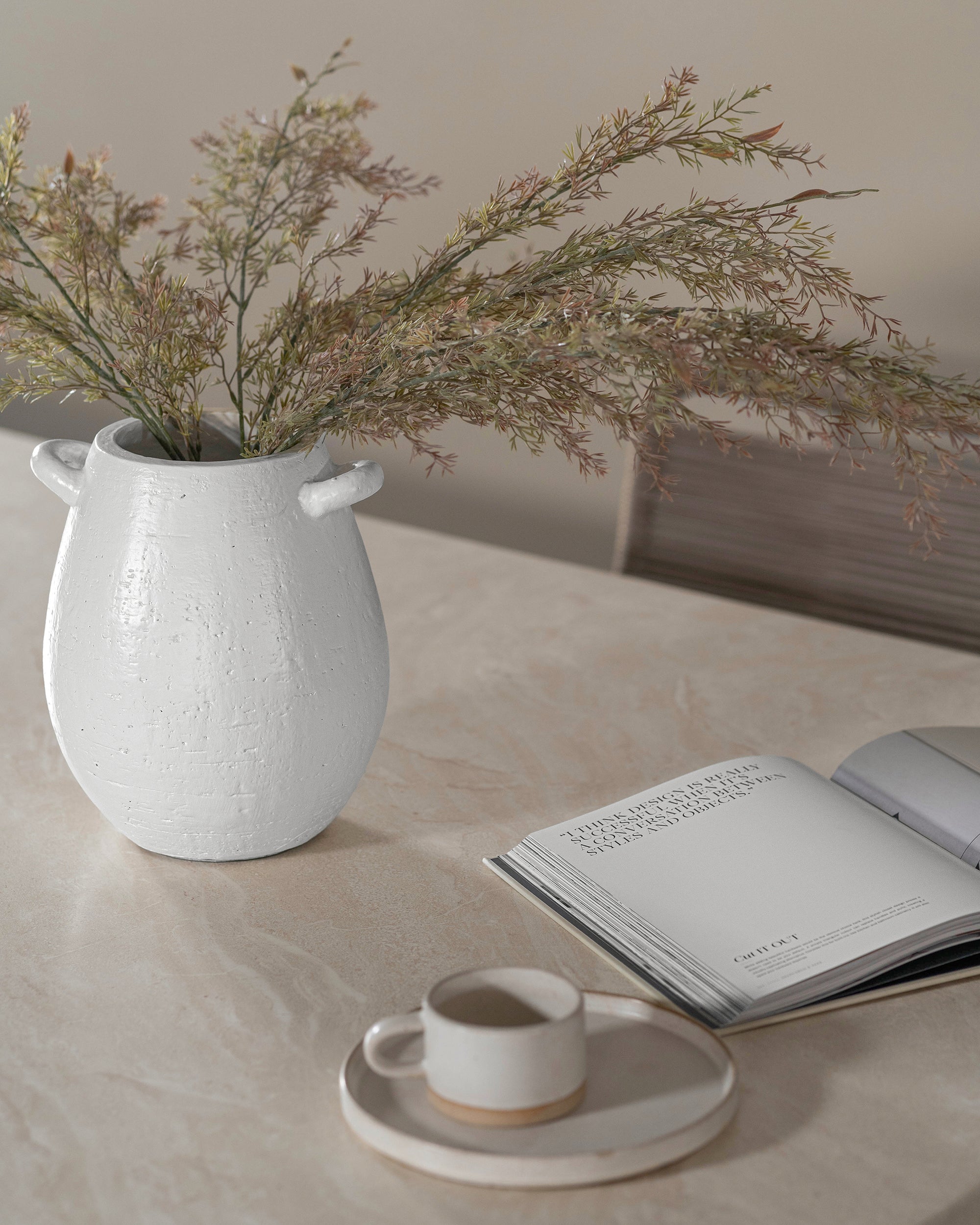 A textured white ceramic vase with dried foliage sits elegantly on a stone table, accompanied by an open design book and a handcrafted ceramic cup on a matching saucer