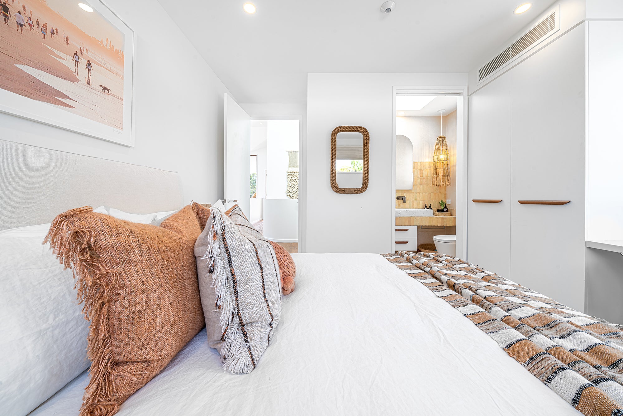 Modern coastal bedroom with neutral-toned bedding, textured cushions, and an ensuite featuring a rattan pendant light and earthy finishes