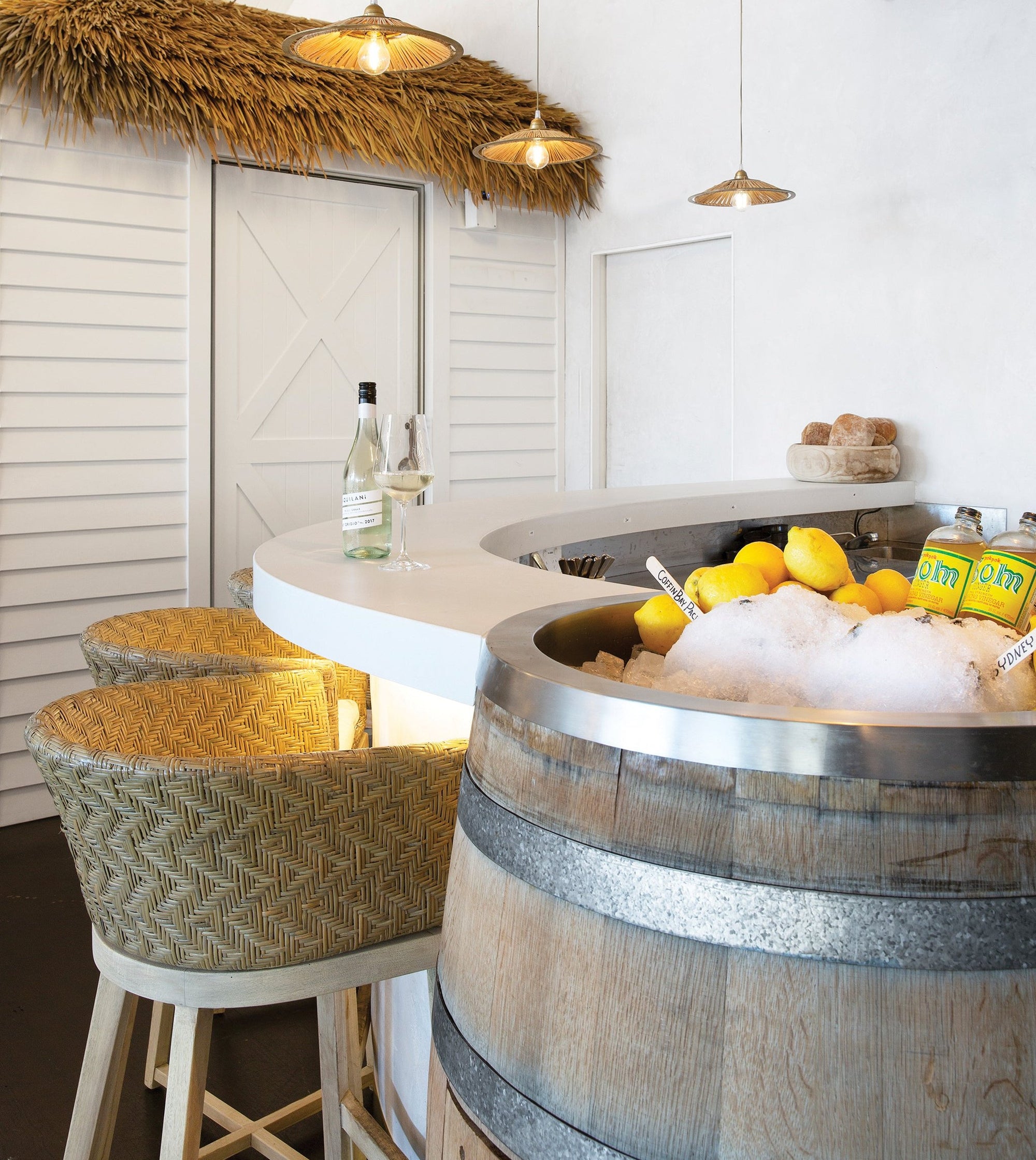 Coastal-style bar setting featuring woven rattan bar stools, a curved white bar counter, thatch pendant lighting, and a rustic barrel ice tub filled with drinks and fresh lemons