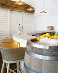 Coastal-style bar setting featuring woven rattan bar stools, a curved white bar counter, thatch pendant lighting, and a rustic barrel ice tub filled with drinks and fresh lemons