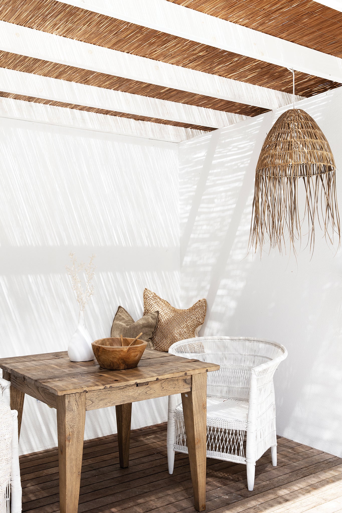 Coastal-inspired outdoor dining space featuring a rustic timber table, a white woven armchair, and a handwoven rattan pendant light. Neutral-toned cushions and natural decor elements enhance the relaxed, sunlit setting beneath a reed pergola.