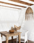 Coastal-inspired outdoor dining space featuring a rustic timber table, a white woven armchair, and a handwoven rattan pendant light. Neutral-toned cushions and natural decor elements enhance the relaxed, sunlit setting beneath a reed pergola.