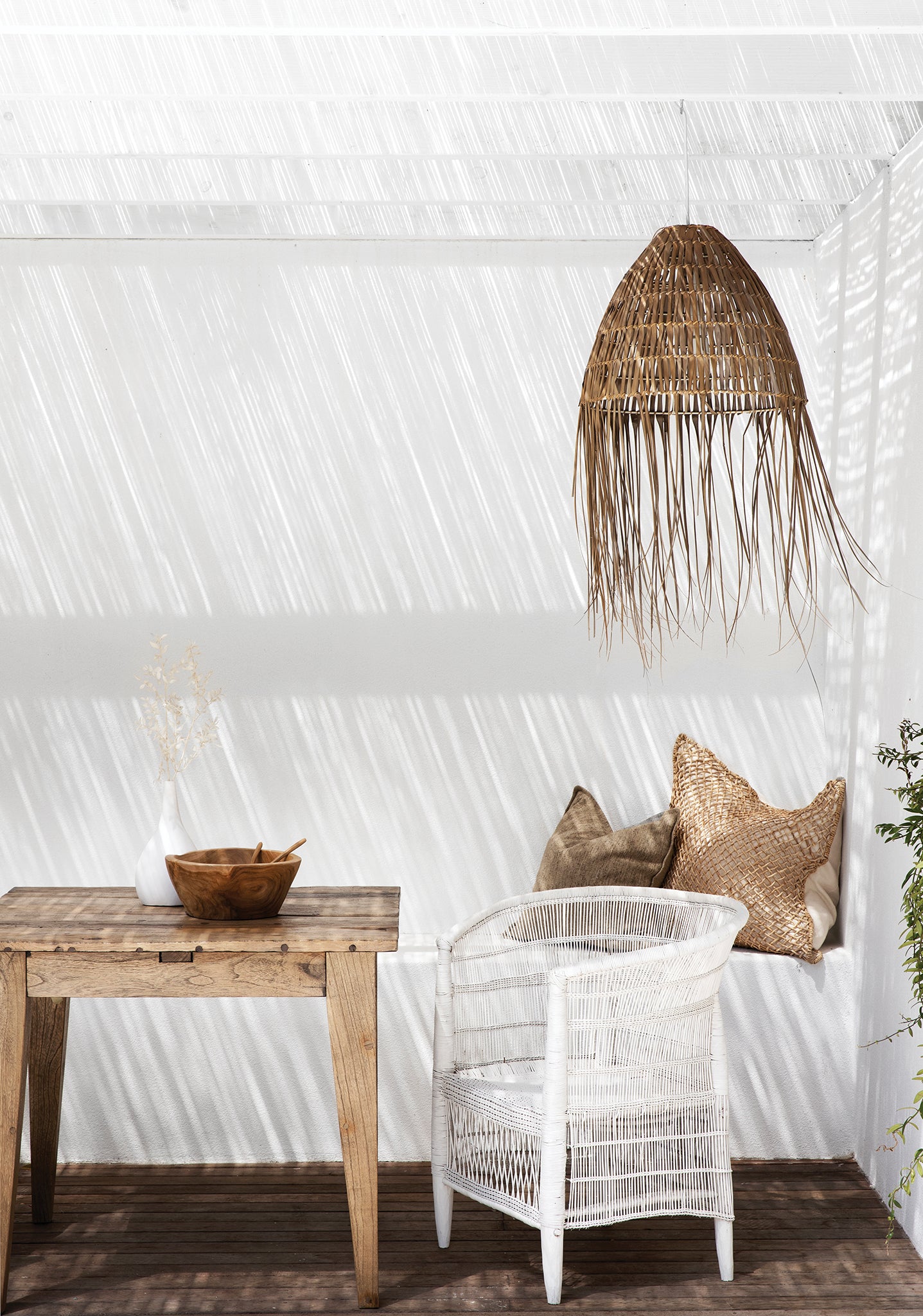 Coastal-inspired outdoor dining space featuring a rustic timber table, a white woven armchair, and a handwoven rattan pendant light. Neutral-toned cushions and natural decor elements enhance the relaxed, sunlit setting beneath a reed pergola.