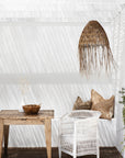 Coastal-inspired outdoor dining space featuring a rustic timber table, a white woven armchair, and a handwoven rattan pendant light. Neutral-toned cushions and natural decor elements enhance the relaxed, sunlit setting beneath a reed pergola.