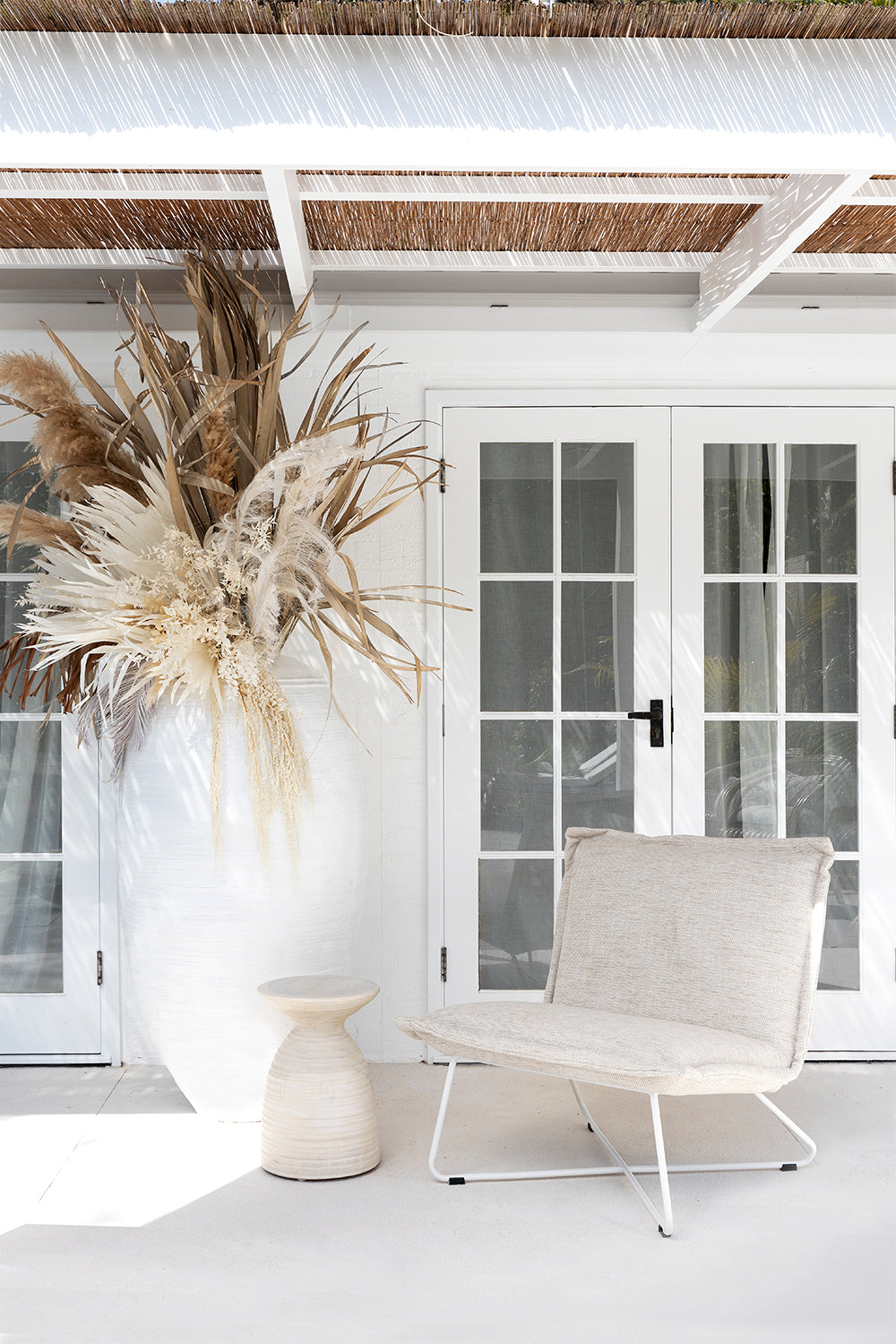 Serene outdoor setting by KULALA Living featuring a neutral-toned lounge chair, textured side table, and oversized ceramic vase with dried floral arrangement, evoking a relaxed coastal aesthetic