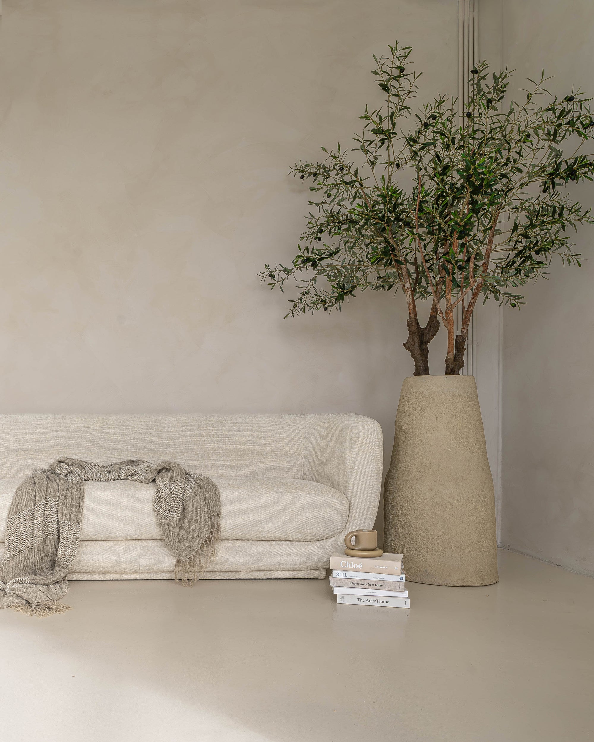 Elegant cream boucle sofa styled with a handwoven throw, ceramic vase, and an olive tree in a sculptural planter, creating a serene neutral-toned living space