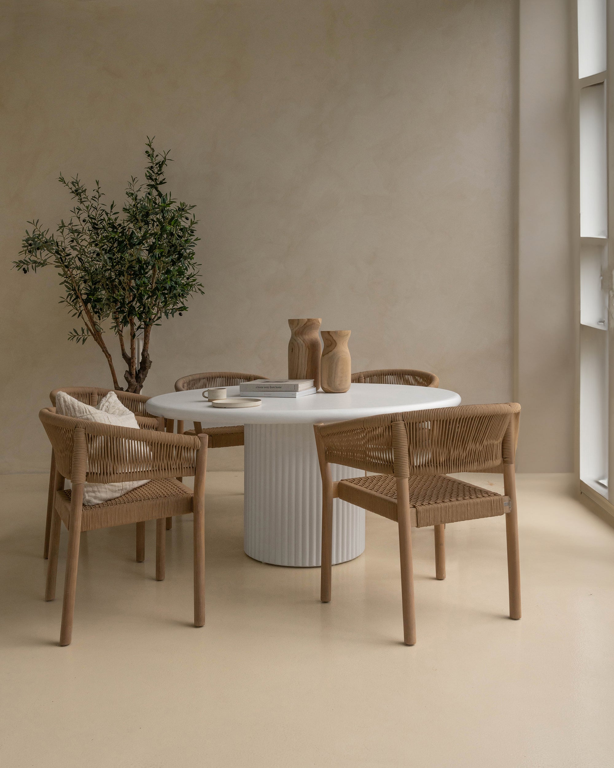 Modern dining space featuring a white fluted round dining table surrounded by woven armchairs in natural timber. Styled with wooden vases, ceramic tableware, and an olive tree, creating a warm, neutral-toned aesthetic.