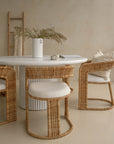 Coastal-inspired dining space featuring curved rattan dining chairs with upholstered seats, paired with a white fluted dining table. Styled with a ceramic vase, neutral-toned decor, and a wooden ladder with a draped throw for a relaxed, organic ambiance