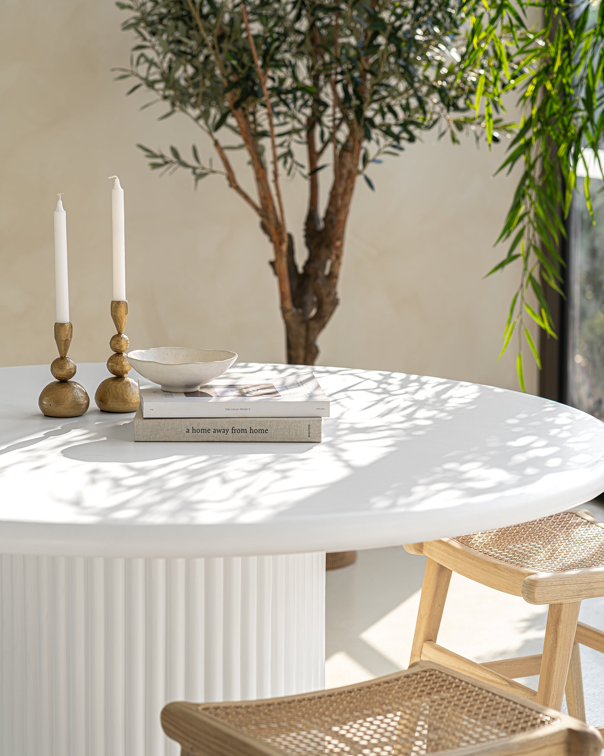 Close-up of a fluted white dining table styled with brass candlesticks, ceramic decor, and stacked books, paired with lightwood cane dining chairs. Sunlight filters through an olive tree, casting soft shadows for a warm, organic ambiance