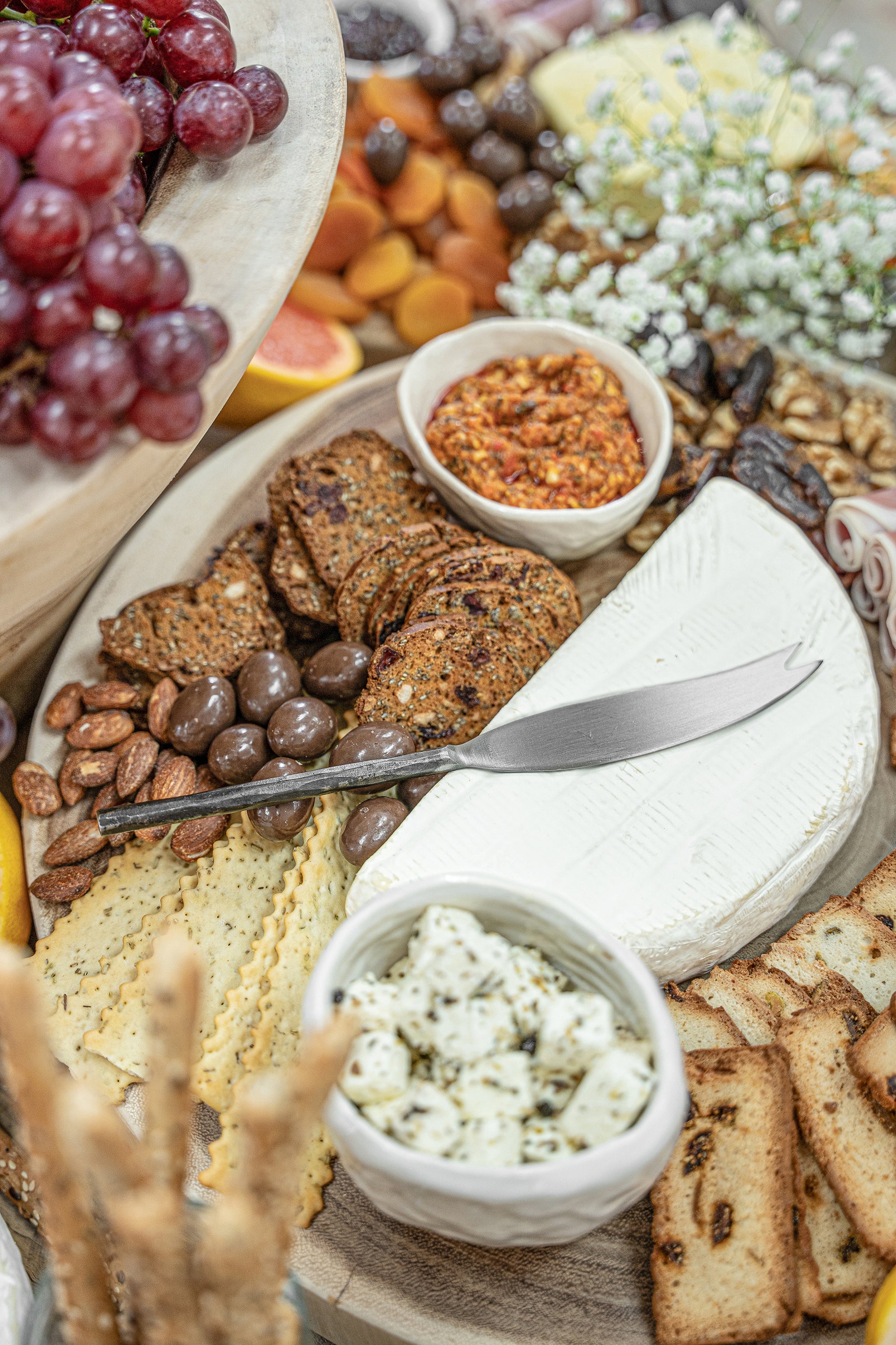 A luxurious grazing board featuring KULALA Living’s hand-forged stainless steel cheese knife resting on a wedge of creamy brie, surrounded by gourmet crackers, marinated feta, almonds, chocolate-coated treats, and vibrant accompaniments. A refined entertaining essential.