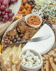 A luxurious grazing board featuring KULALA Living’s hand-forged stainless steel cheese knife resting on a wedge of creamy brie, surrounded by gourmet crackers, marinated feta, almonds, chocolate-coated treats, and vibrant accompaniments. A refined entertaining essential.