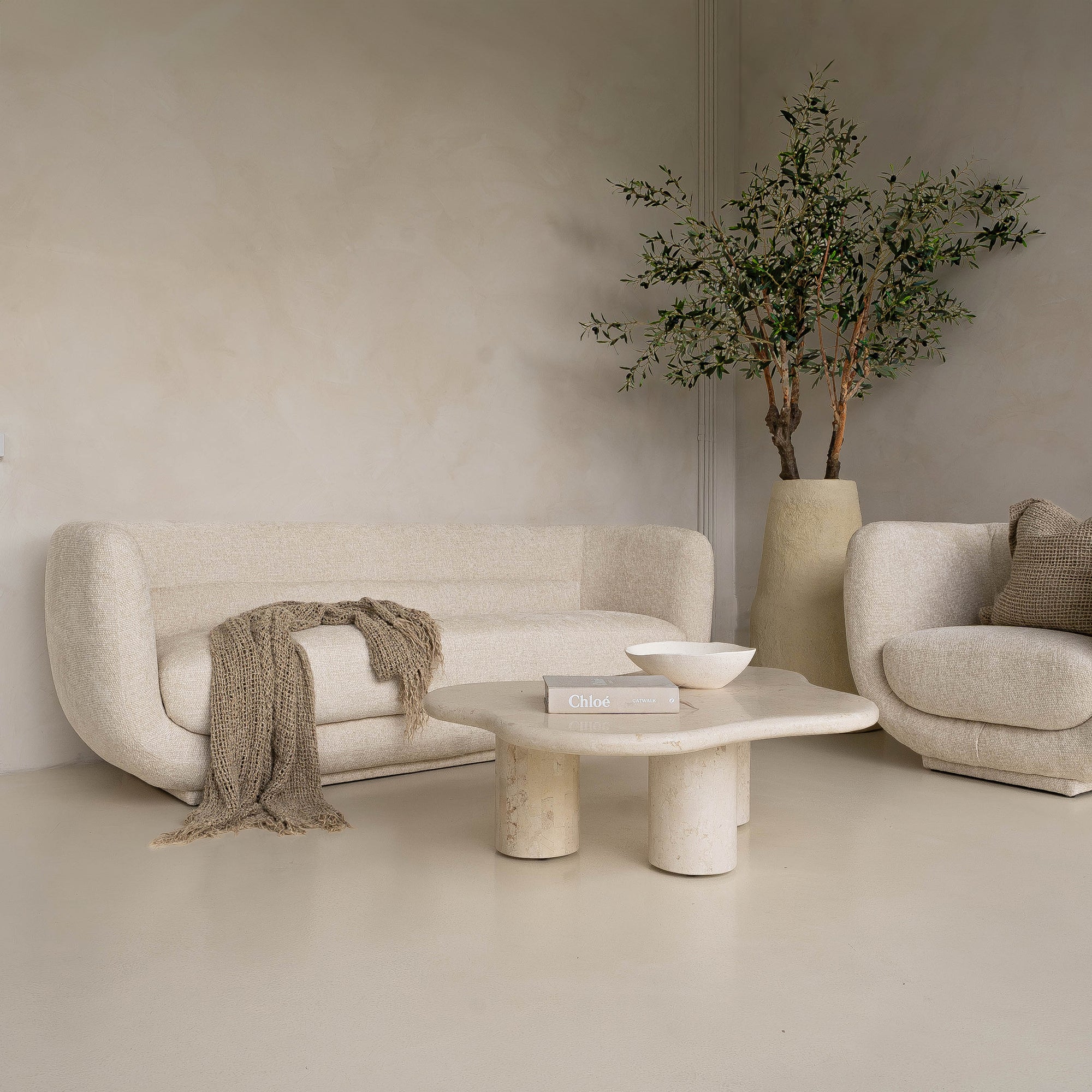 A modern, minimalist lounge setting featuring the KULALA Living curved bouclé sofa and matching armchair in a soft ivory hue. Styled with a textured throw, linen cushions, a sculptural stone coffee table, and a statement vase with an olive tree for a tranquil, contemporary aesthetic