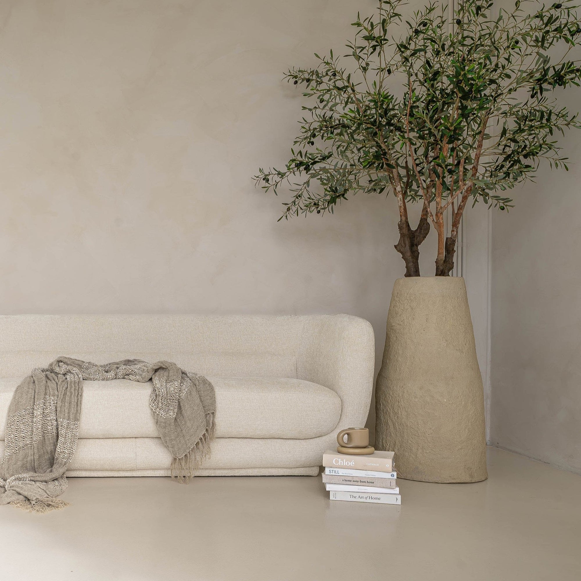 A modern, minimalist lounge setting featuring the KULALA Living curved bouclé sofa and matching armchair in a soft ivory hue. Styled with a textured throw, linen cushions, a sculptural stone coffee table, and a statement vase with an olive tree for a tranquil, contemporary aesthetic