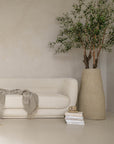 A modern, minimalist lounge setting featuring the KULALA Living curved bouclé sofa and matching armchair in a soft ivory hue. Styled with a textured throw, linen cushions, a sculptural stone coffee table, and a statement vase with an olive tree for a tranquil, contemporary aesthetic