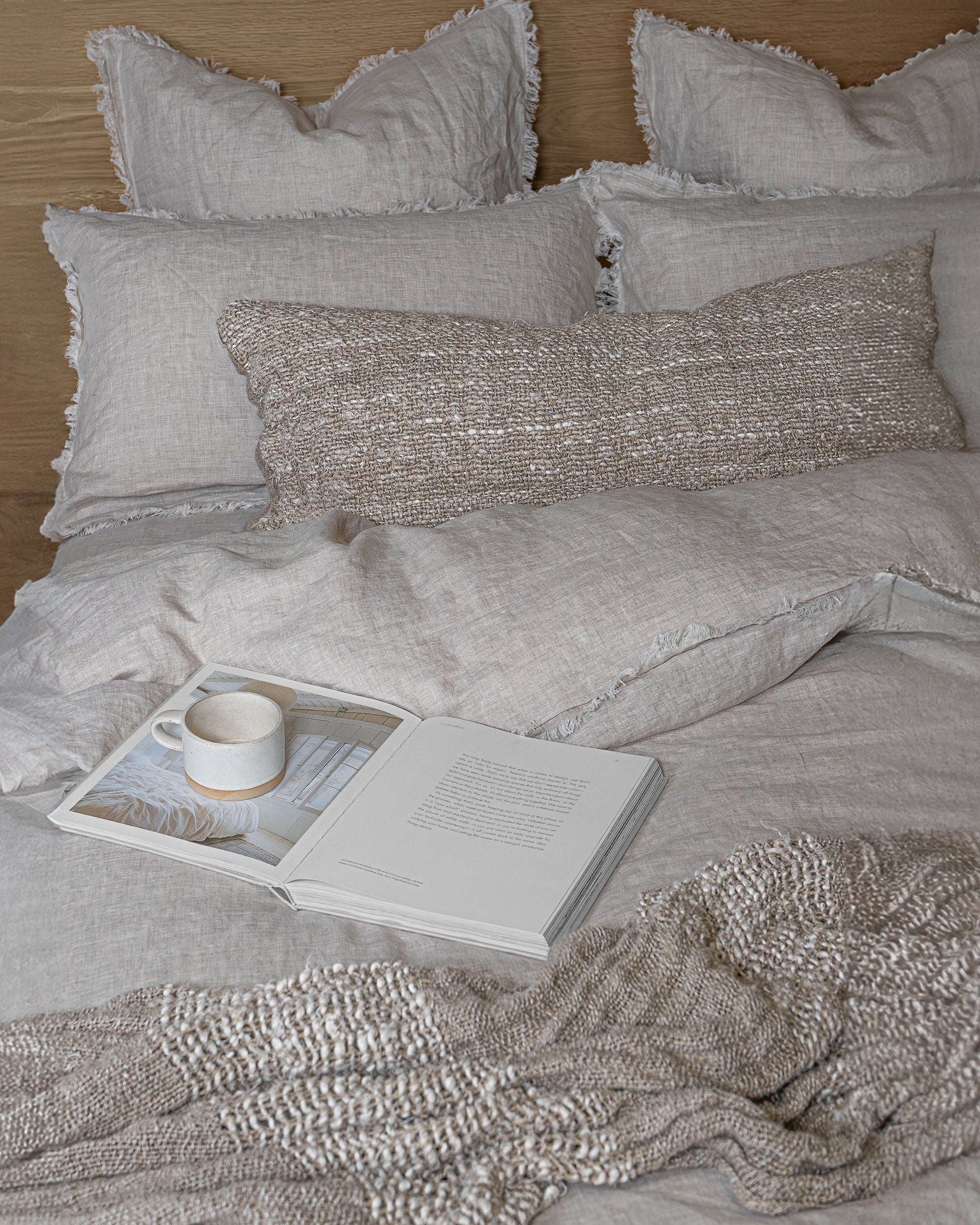 Serene bedroom setting featuring soft fringed linen bedding, a textured handwoven cushion, and a cosy throw, styled with an open book and ceramic mug for a relaxed atmosphere