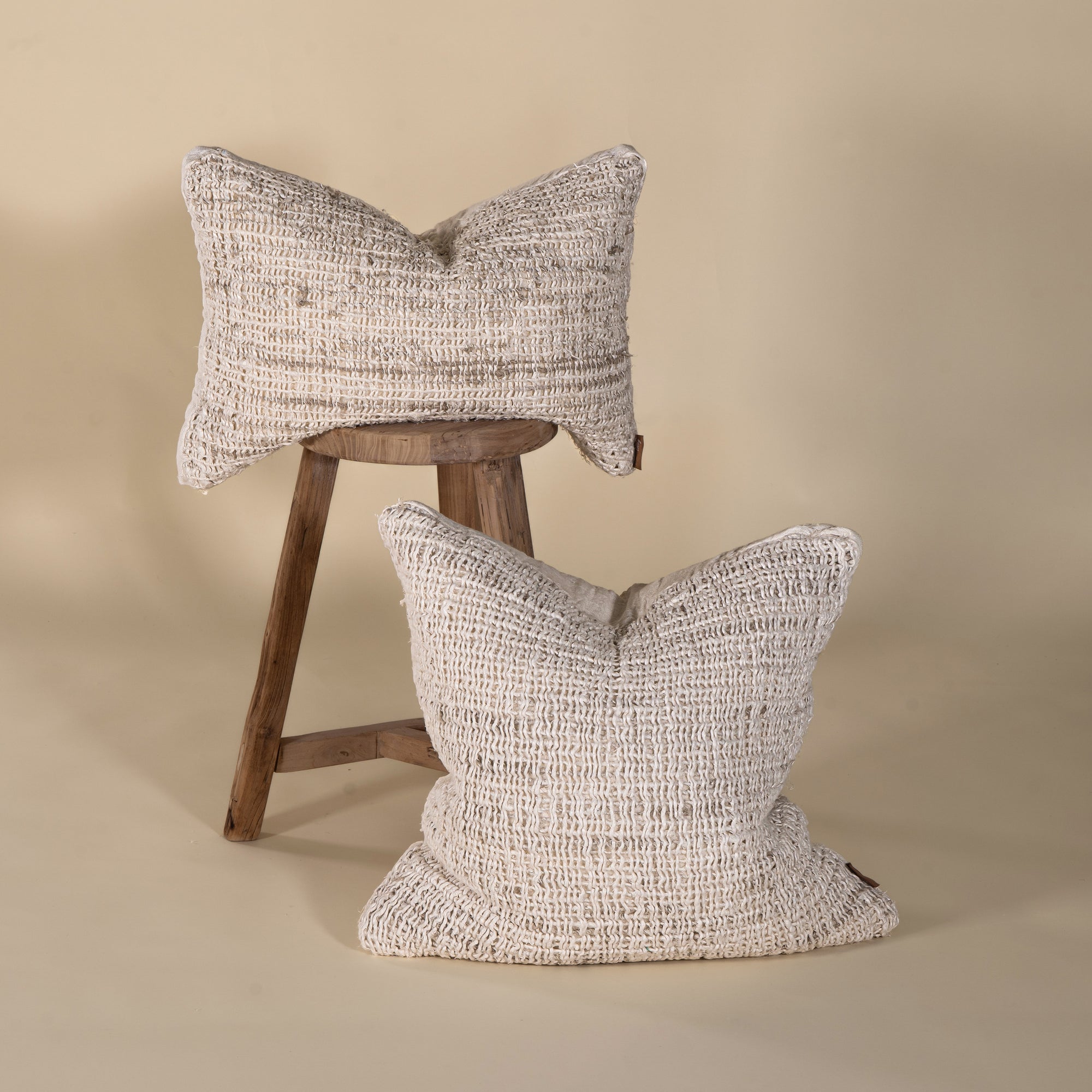 Set of two handwoven cushions in a neutral tone, showcasing intricate texture and artisanal craftsmanship, styled with a rustic wooden stool for a relaxed, organic feel