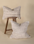 Set of two handwoven cushions in a neutral tone, showcasing intricate texture and artisanal craftsmanship, styled with a rustic wooden stool for a relaxed, organic feel