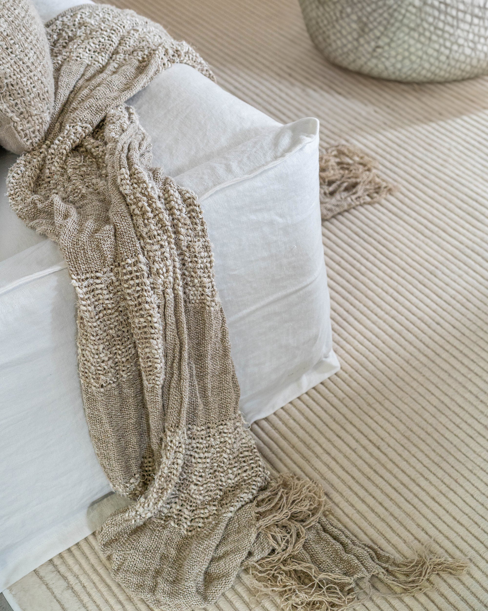 Handwoven fringed throw in a soft neutral hue, draped over a white linen sofa, adding texture and warmth to a serene, minimalist living space