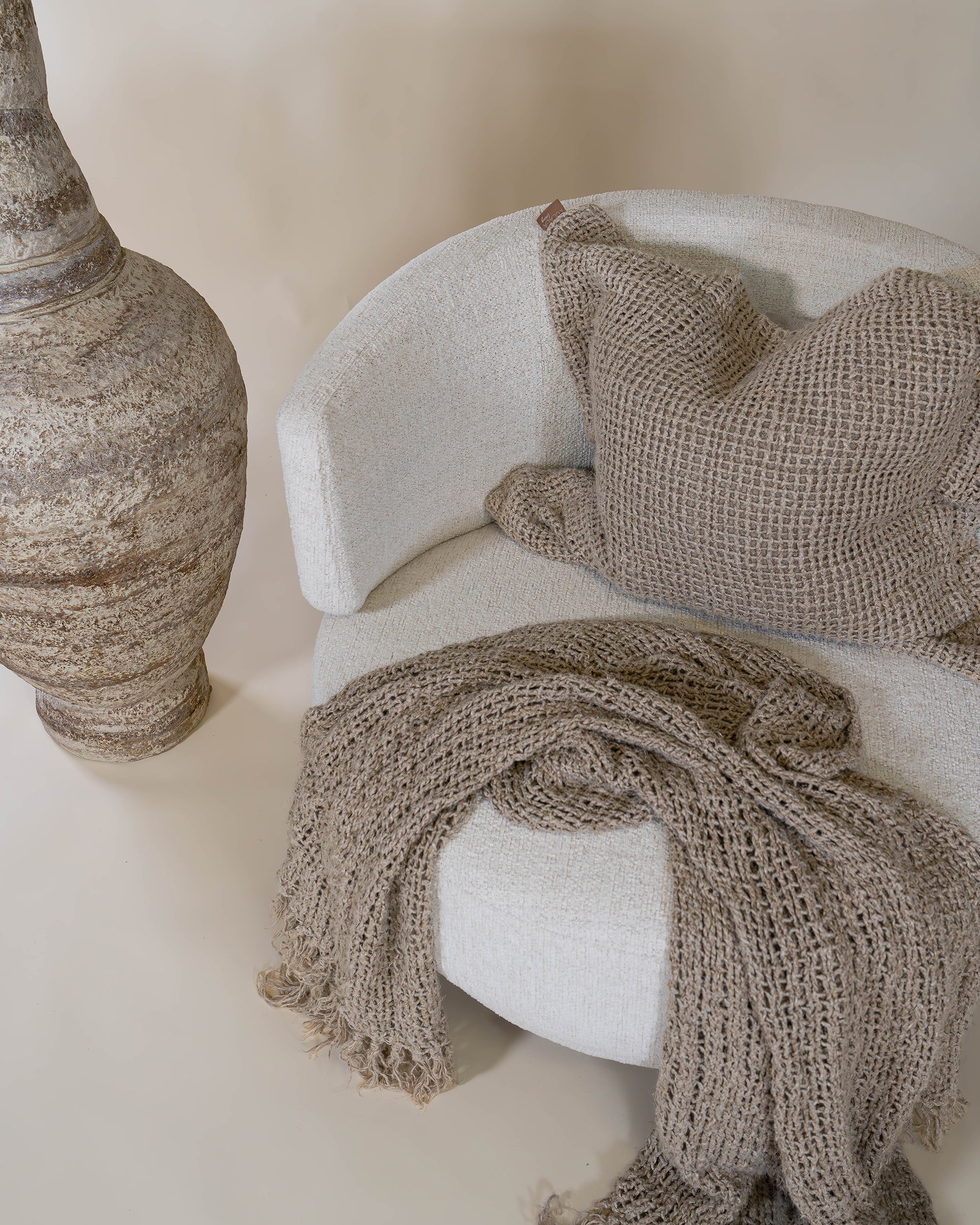 A cosy handwoven taupe throw blanket and matching cushion styled on a curved cream occasional chair, creating a warm and inviting space